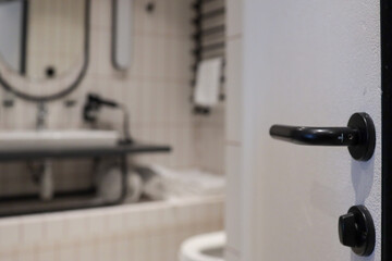 Close-focus shot of black door handle with modern hotel bathroom softly blurred behind—oval mirror, washbasin and towels—conveying clean contemporary design