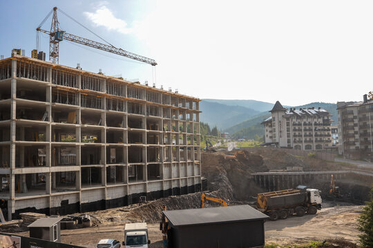 Concrete skeleton of multi-storey building rises beside a tower crane on busy mountain-resort construction site; dump truck and excavators dig foundations against a clear summer sky