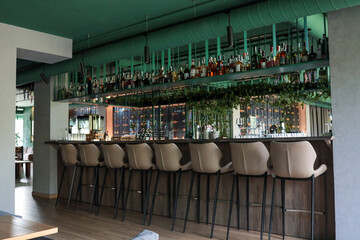 Stylish hotel cocktail bar with long wooden counter, sleek beige stools, suspended greenery and...