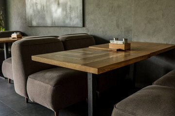 Low brown upholstered sofas and oak tables line a minimalist café lounge with grey walls, forest artwork and soft daylight, creating a calm, modern seating atmosphere