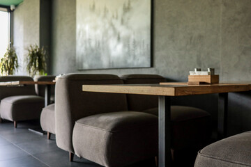 Low brown upholstered sofas and oak tables line a minimalist café lounge with grey walls, forest artwork and soft daylight, creating a calm, modern seating atmosphere