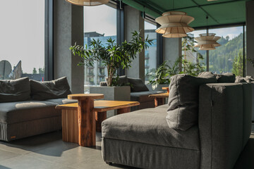 Sun-lit hotel lounge with panoramic windows, grey modular sofas, rustic oak tables, sculptural...