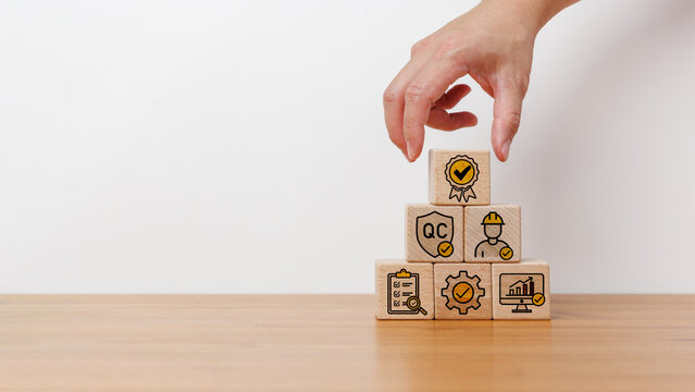 Business concept of quality assurance and control. Hand arranging wooden blocks with quality control, inspection, and performance icons in pyramid shape on wooden table in white background.
