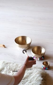 Three Tibetan singing bowls placed on a wooden floor, accompanied by colorful pads and mallets. A hand holds a mallet, ready to play the soothing, resonant tones of the bowls