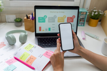 Person using smartphone at home office desk with laptop and marketing strategy on screen