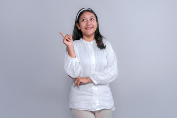 Young Asian woman in white blouse smiling and pointing upward with index finger, looking curious...