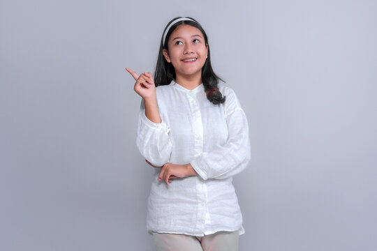 Young Asian woman in white blouse smiling and pointing upward with index finger, looking curious and inspired. Studio portrait on grey background, ideal for creative campaign visuals. - Powered by Adobe