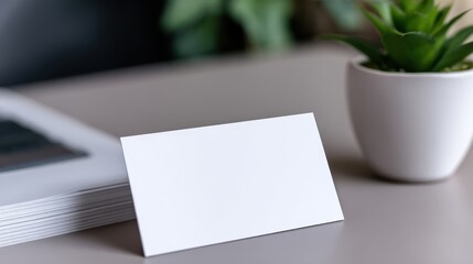 Minimalist workspace featuring a blank card on a desk with a plant and laptop in the background