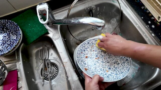 Housekeeper scrubbing dirty dishes in the kitchen sink with yellow sponge