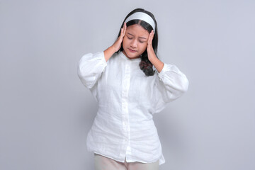 Frustrated young Asian woman in white outfit standing with eyes closed and hands on her temples, expressing stress or headache. Studio shot with gray background, copy space.