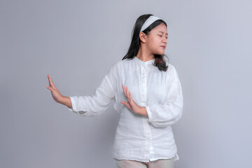Asian woman in white clothes making a hand gesture to reject something while turning her face away. Her closed eyes and serious expression emphasize disapproval, isolated on gray background.
