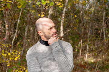 Pensive bald man outdoors in autumn, hand on chin, looking sideways, lost in thought. Grey sweater,...