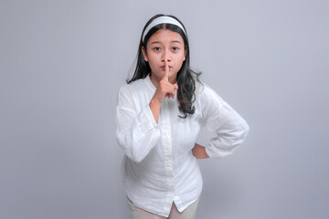 Portrait of a serious Asian woman in white outfit making a “shh” gesture with index finger on lips, signaling silence or secrecy, isolated in studio with gray background.
