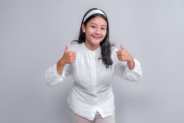 Happy young Asian woman with long black hair wearing a white shirt and headband, leaning forward while smiling and showing a double thumbs up gesture, standing in a bright studio setting.