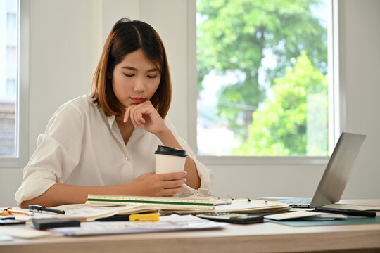 Young female engineer in a white shirt holding a coffee cup, thoughtfully looking at her laptop in a bright modern office