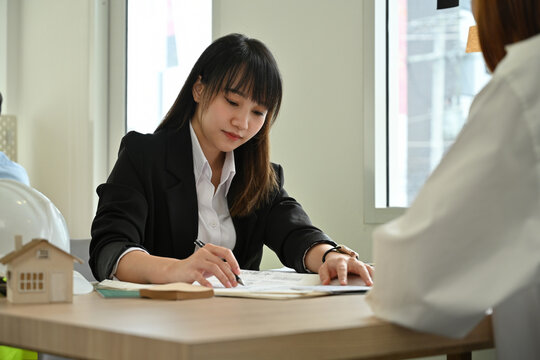 Young female architect in a black blazer studying architectural plans at her desk in a bright modern office.