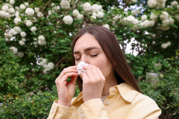 Woman with paper tissue suffering from seasonal pollen allergy near blossoming shrub on spring day