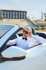 Mature man in bow tie and sunglasses enjoying ride in white cabriolet.