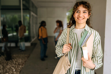College student smiles while holding books and standing in university courtyard during sunny day