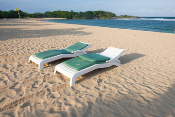 Two empty beach lounge chairs with green cushions sit on golden sand near the ocean shore under...