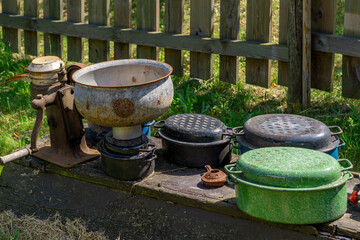 Set of old kitchen utensils displayed outdoors by a wooden fence — including cast iron pots, a...