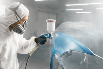 Car repair painter in protective overalls and painting a respirator car wing in the paint chamber