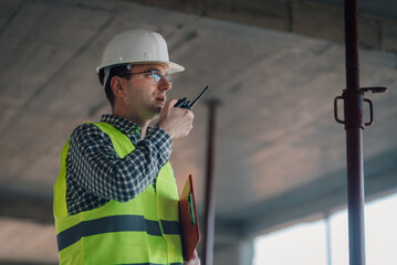 Construction engineer using walkie talkie and holding clipboard inside building under construction