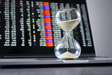 Close-up of glass hourglass standing on laptop, stock market quotes on screen in background.