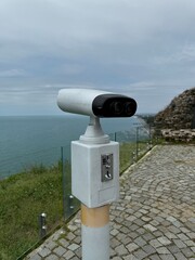 Coin-operated binocular viewer mounted on a pole at a coastal viewpoint with a glass barrier and...