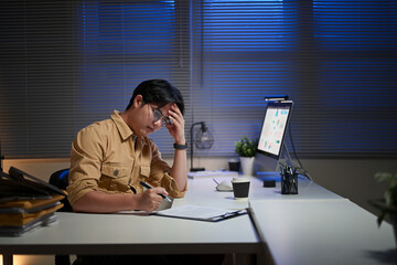 Tired employee holding his head in frustration while reviewing documents late at night in an office