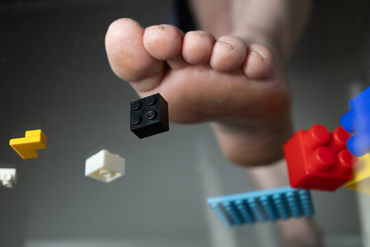 A foot is shown stepping on colorful construction toy bricks scattered on the floor, highlighting the chaos of playtime. The setting suggests a fun but messy environment created by children.