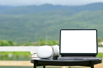 A laptop is on a table with headphones next to it. The laptop is white screen. The scene is set in a natural environment, with mountains in the background