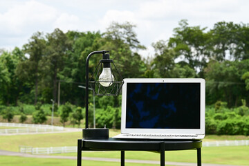 A laptop is on a table next to a lamp. The lamp is turned off, and the laptop screen is black. The scene is set in a grassy field, with trees in the background