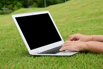 A person is typing on a laptop in a grassy field. The laptop is open and the screen is black