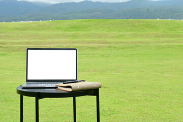 A laptop is open on a table in a grassy field. The grass is green and the sky is cloudy. The scene is peaceful and serene, with the laptop