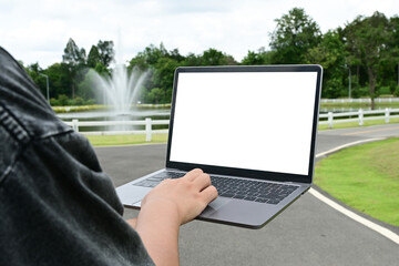 A person is holding a laptop in a park. The laptop screen is blank, and the person is typing on it.
