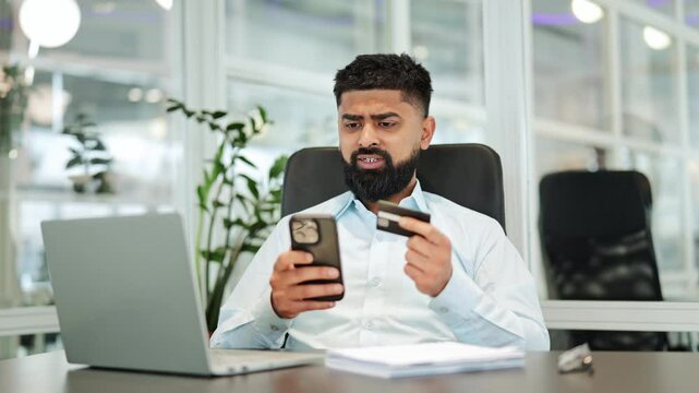 Surprised handsome Indian businessman wearing a light shirt experiences online payment issue using smartphone and credit card in modern office, showing frustration and confusion.