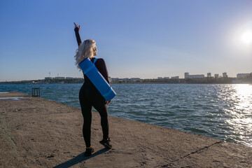 Yoga Woman Lake - A woman in black leggings with a blue yoga mat over her shoulder, standing on a pier overlooking a lake, pointing towards the sun.