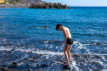 Teen boy playing in ocean waves on black volcanic sand beach, joyful summer moment, natural fun,...