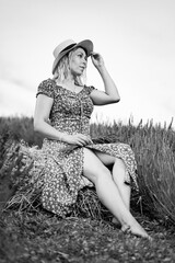 Woman in floral dress sits on hay bale in field, enjoying summer day.