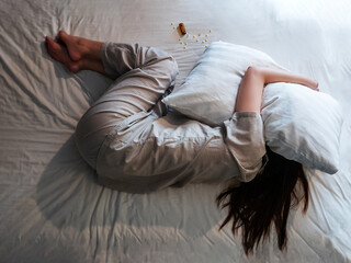 woman lying on bed, cover her head with pillow suffer from insomnia, sleeping pills next to her on bed. top above view of woman curl up in stress in bedroom with sleep disorder