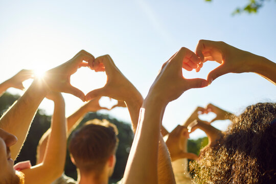 Group of young adult youth people doing heart gesture against bright blue sunny sky. Friends hold up hands making heart shapes in sunlight feeling love and connection. Love, friendship, nature concept