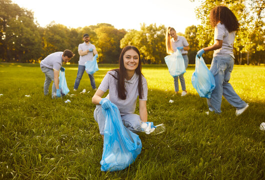 Portrait of a happy young woman with gloves and trash bag collecting plastic garbage in the summer park with a people volunteers on the background. Environmental pollution and ecology concept. - Powered by Adobe