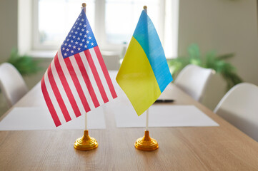 American and Ukrainian flags standing side by side on wooden table in meeting room. Symbol of...
