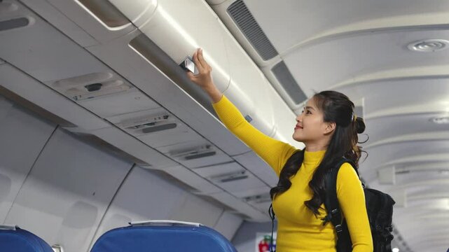 Woman storing travel luggage in overhead bin while boarding airplane, wearing yellow shirt and black backpack against blue cabin interior