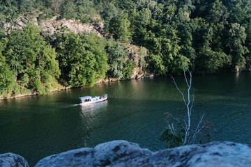 A small white boat cruises on a tranquil river, surrounded by lush green cliffs and dense forests.