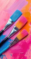 Three colorful paintbrushes resting on a vibrant, abstract background of mixed pink, purple, and orange paint strokes
