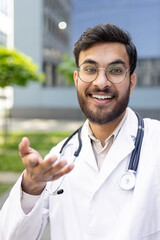 A smiling doctor with a stethoscope gestures towards the viewer while talking on a video call...