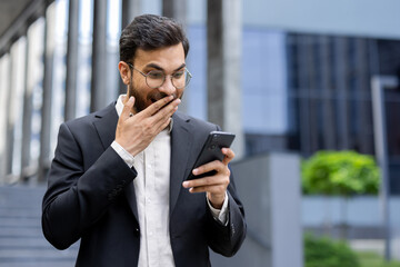 A surprised businessman in a suit looks at his phone with an open mouth, covering it with his hand.