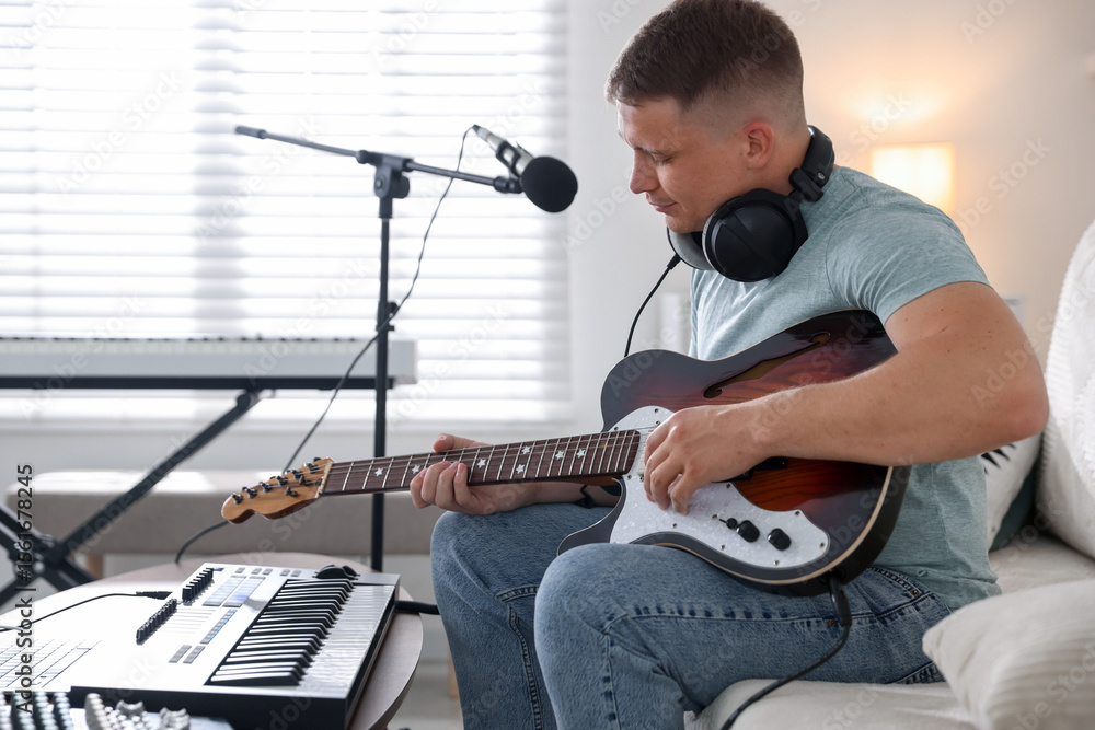 Poster man working with guitar and midi keyboard on sofa in home studio